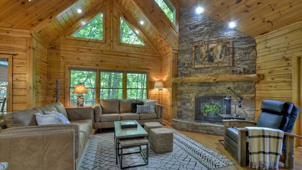 A log cabin interior with a stone fireplace, wooden furniture, and large windows. - Cedar Ridge | Hot Tub + Game Room + Fire Pit (Blue Ridge)