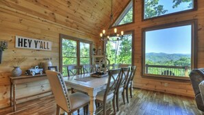 L’intérieur d’un chalet en bois comprend une table à manger, des chaises, un lustre et de grandes fenêtres qui offrent une vue sur l’extérieur.