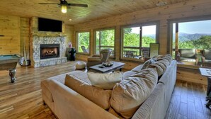 Un intérieur de chalet chaleureux avec une cheminée en pierre, un plancher en bois, un grand canapé et un balcon offrant une vue panoramique.
