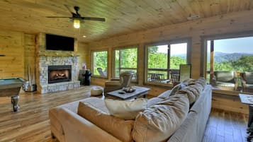 A cozy cabin interior with a stone fireplace, wooden floors, a large sofa, and a balcony with a scenic view.
