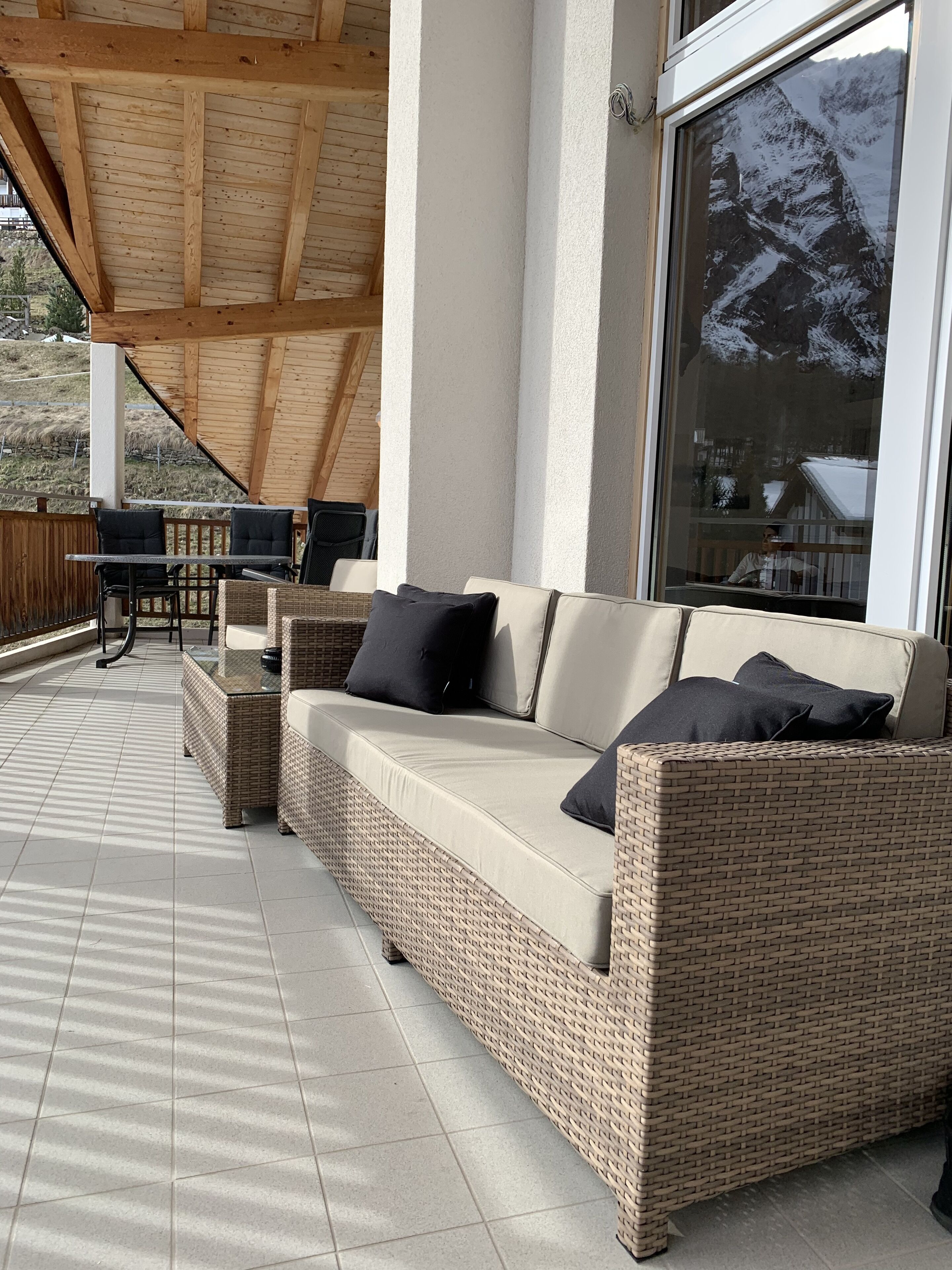 A covered outdoor seating area with a wicker sofa and chairs, a tiled floor, and a view of a snowy mountain.