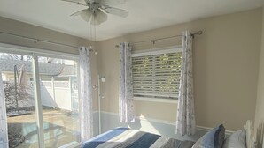 Une chambre avec un lit, une table de chevet, une lampe, un ventilateur de plafond et une vue sur le jardin à travers une porte-fenêtre coulissante.