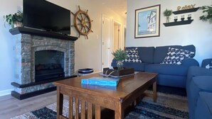 Un salon doté d’une cheminée en pierre, d’un téléviseur à écran plat fixé au-dessus, d’une table basse en bois, d’un canapé bleu agrémenté de coussins à motifs et d’un tapis.