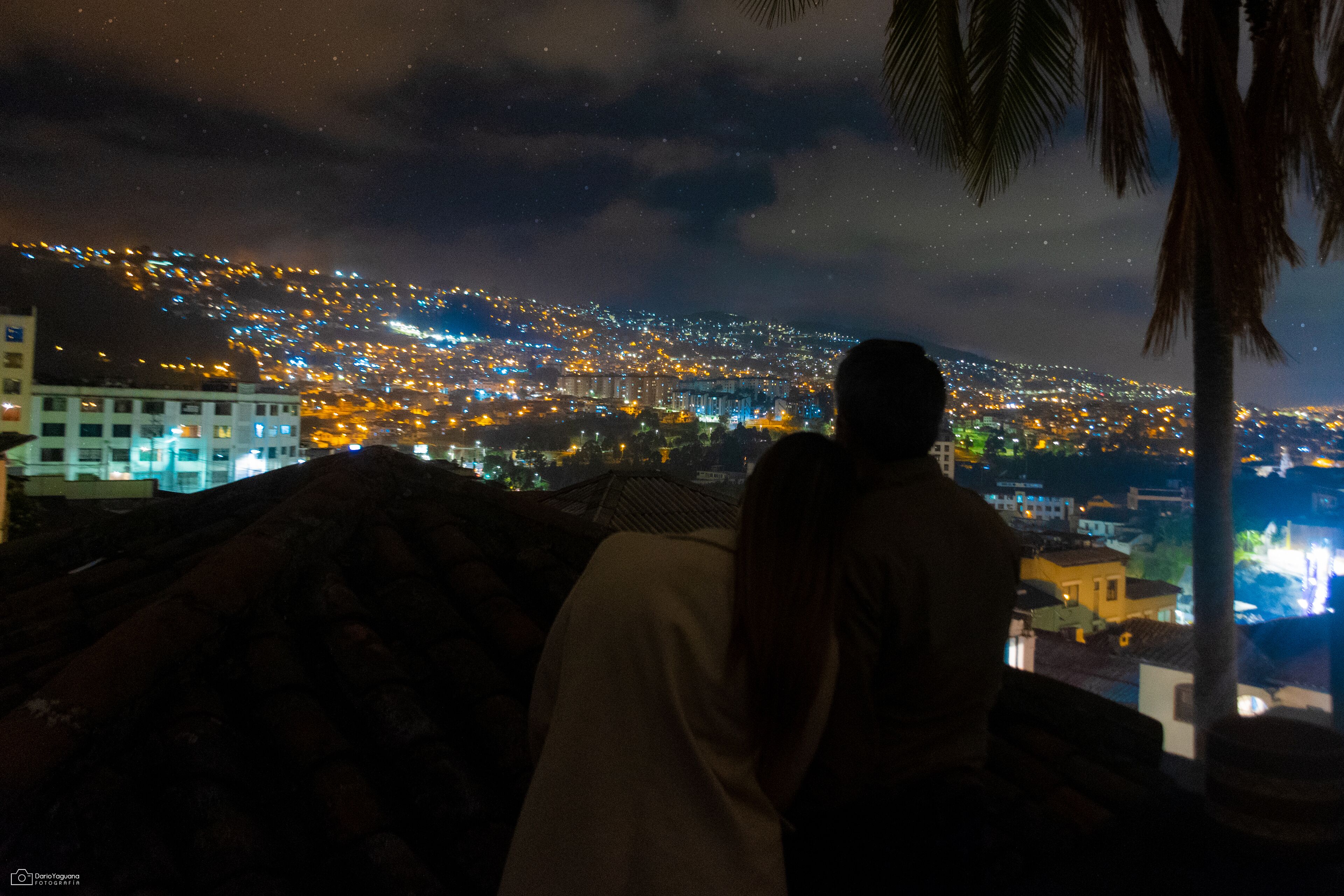 Vista nocturna d'un paisatge urbà amb edificis il·luminats i una persona alçada en un turó observant la ciutat.