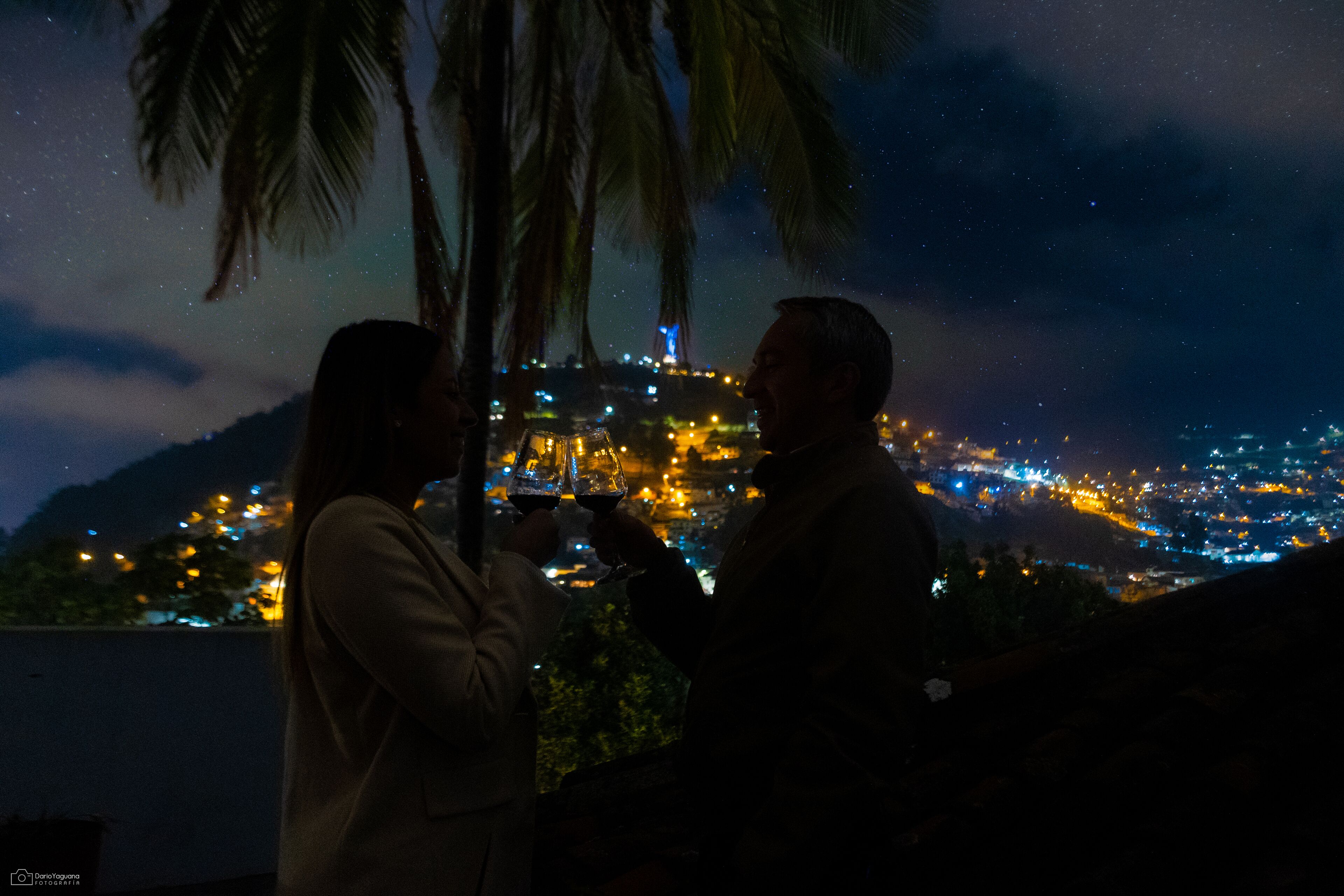 Duas pessoas segurando copos de vinho, com vista para uma paisagem urbana com edifícios iluminados e palmeiras.