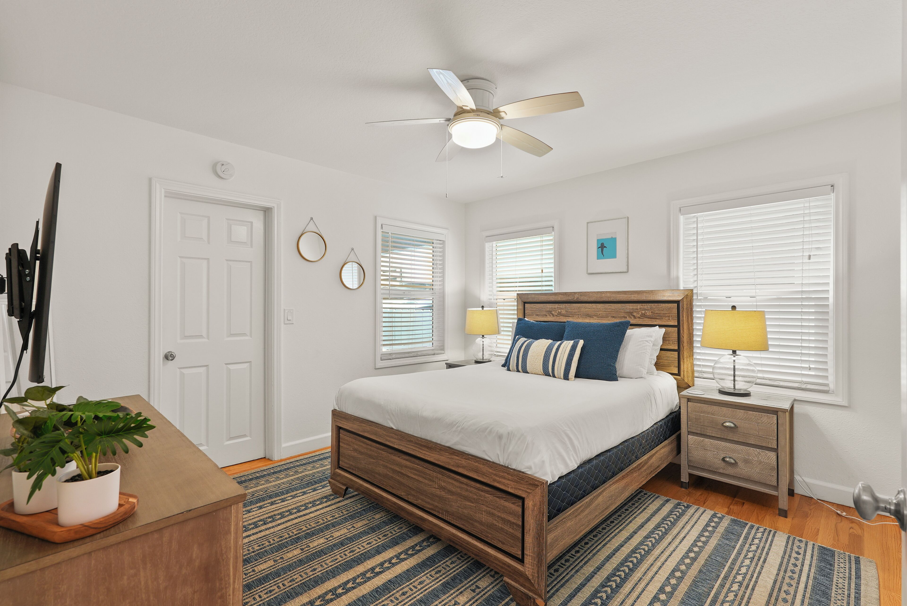 A bedroom with a wooden bed, a ceiling fan, a bedside table with lamps, and a potted plant.