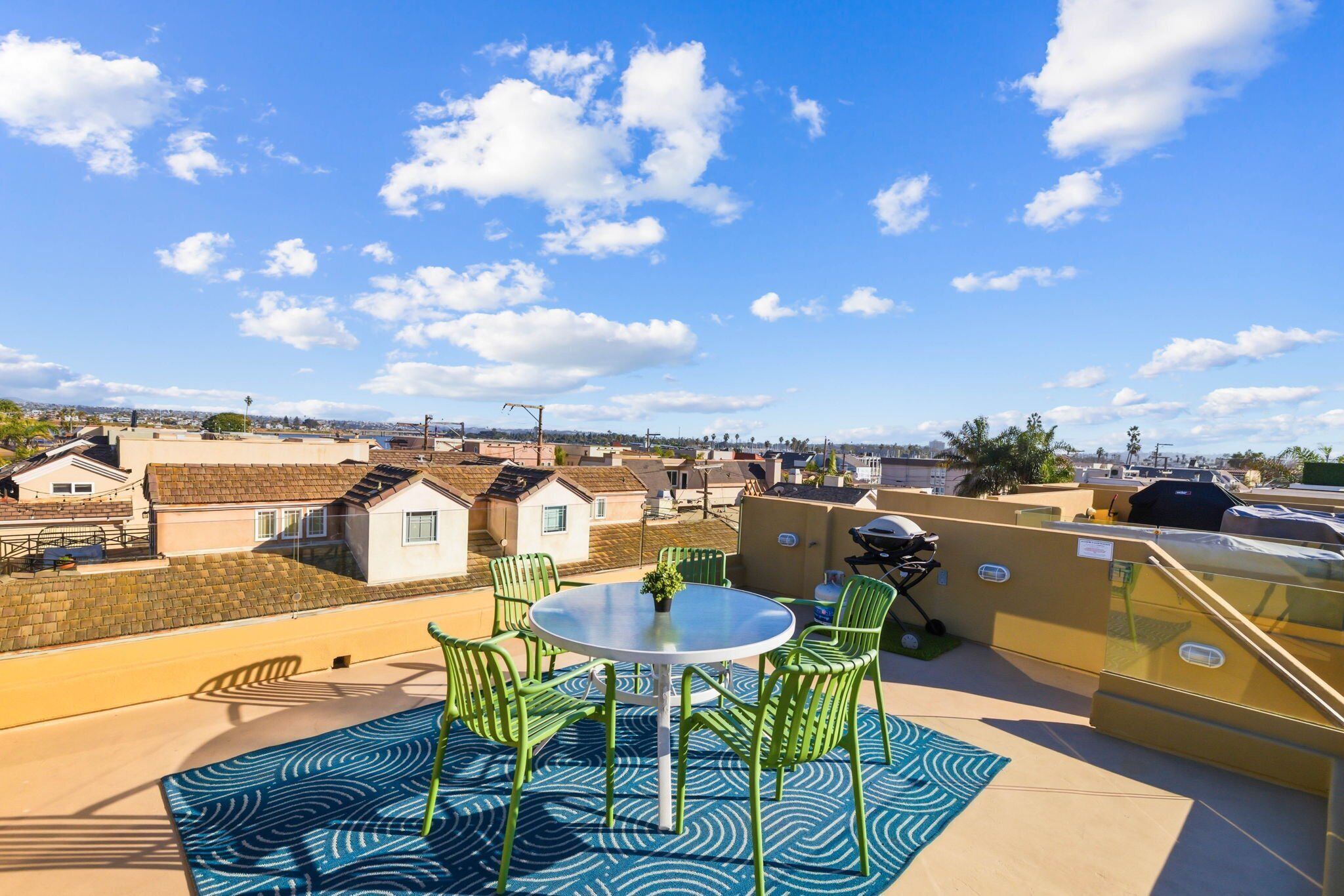 A rooftop terrace with a round table and chairs, a small potted plant, and a view of a residential area.