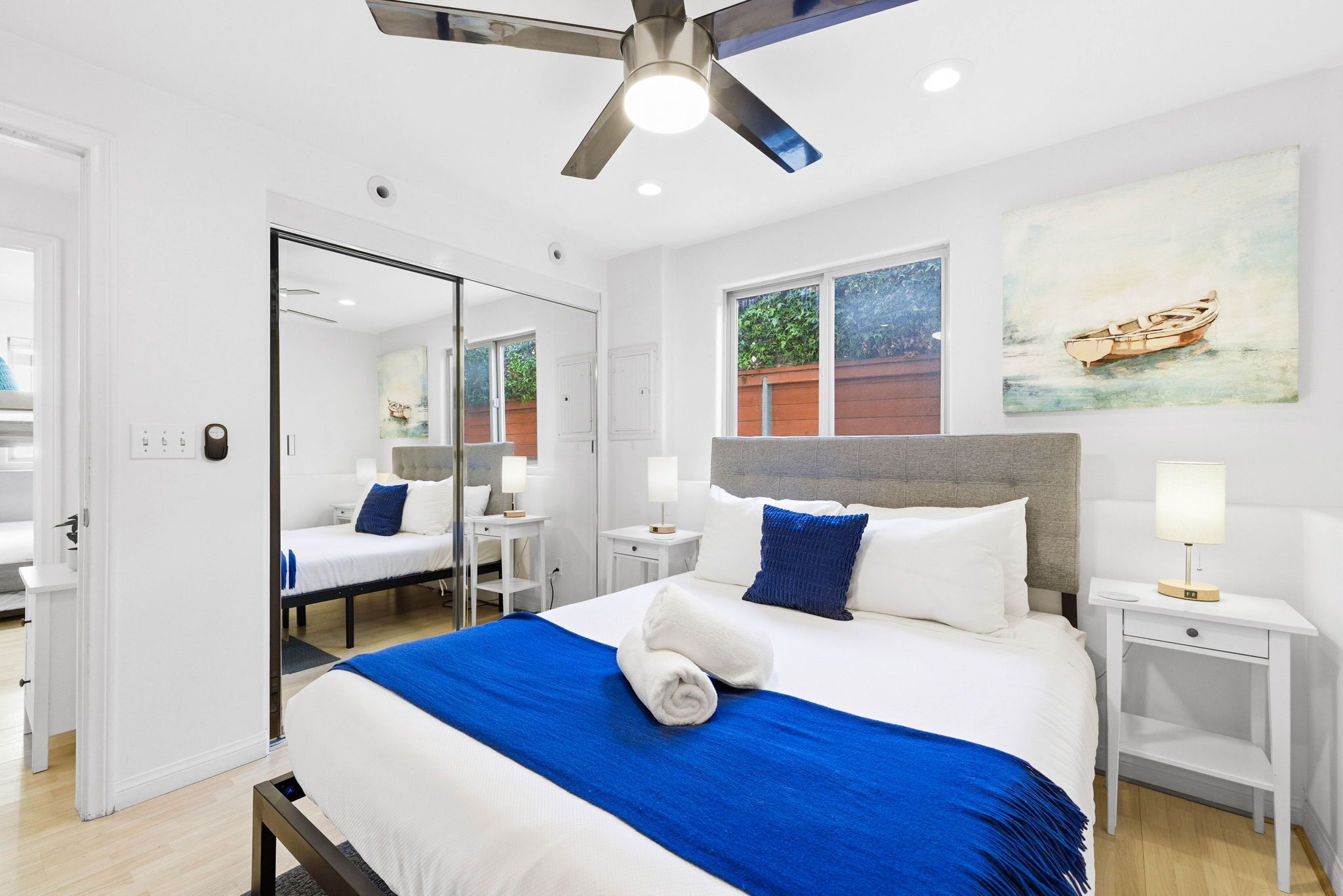 Une chambre d’hôtel moderne avec un grand lit, un ventilateur de plafond, une fenêtre donnant sur un paysage verdoyant et un tableau représentant un bateau accroché au mur.