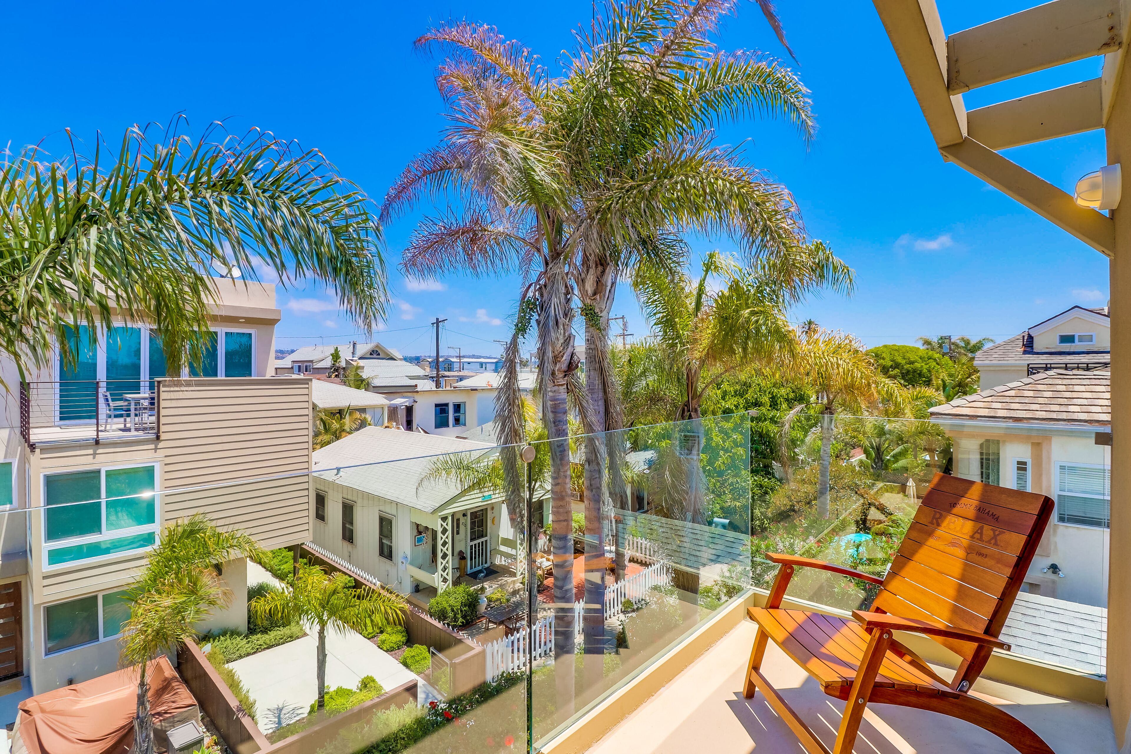 A balcony with a wooden chair, overlooking a residential area with houses and palm trees.