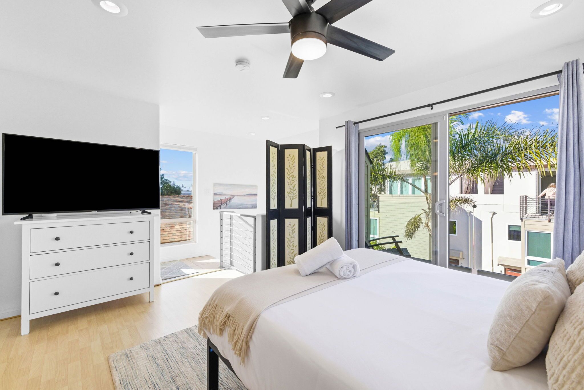 Ein modernes Schlafzimmer mit Flachbildfernseher, Deckenventilator, einem großen Bett mit Ausblick und einer Glasschiebetür, die auf einen Balkon führt.