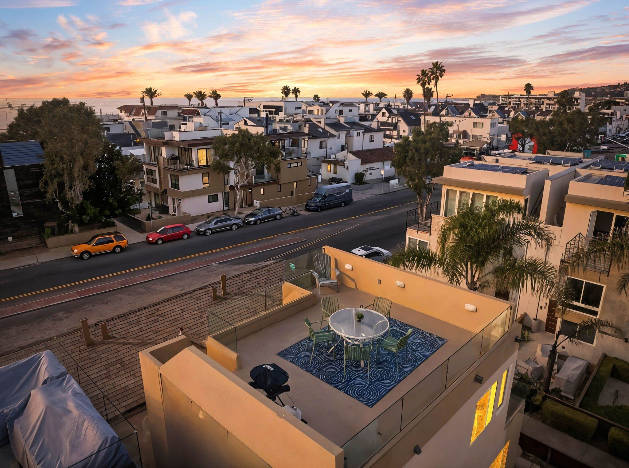 Un barrio residencial con casas modernas, una calle con coches aparcados y una terraza en la azotea con mesa y sillas.