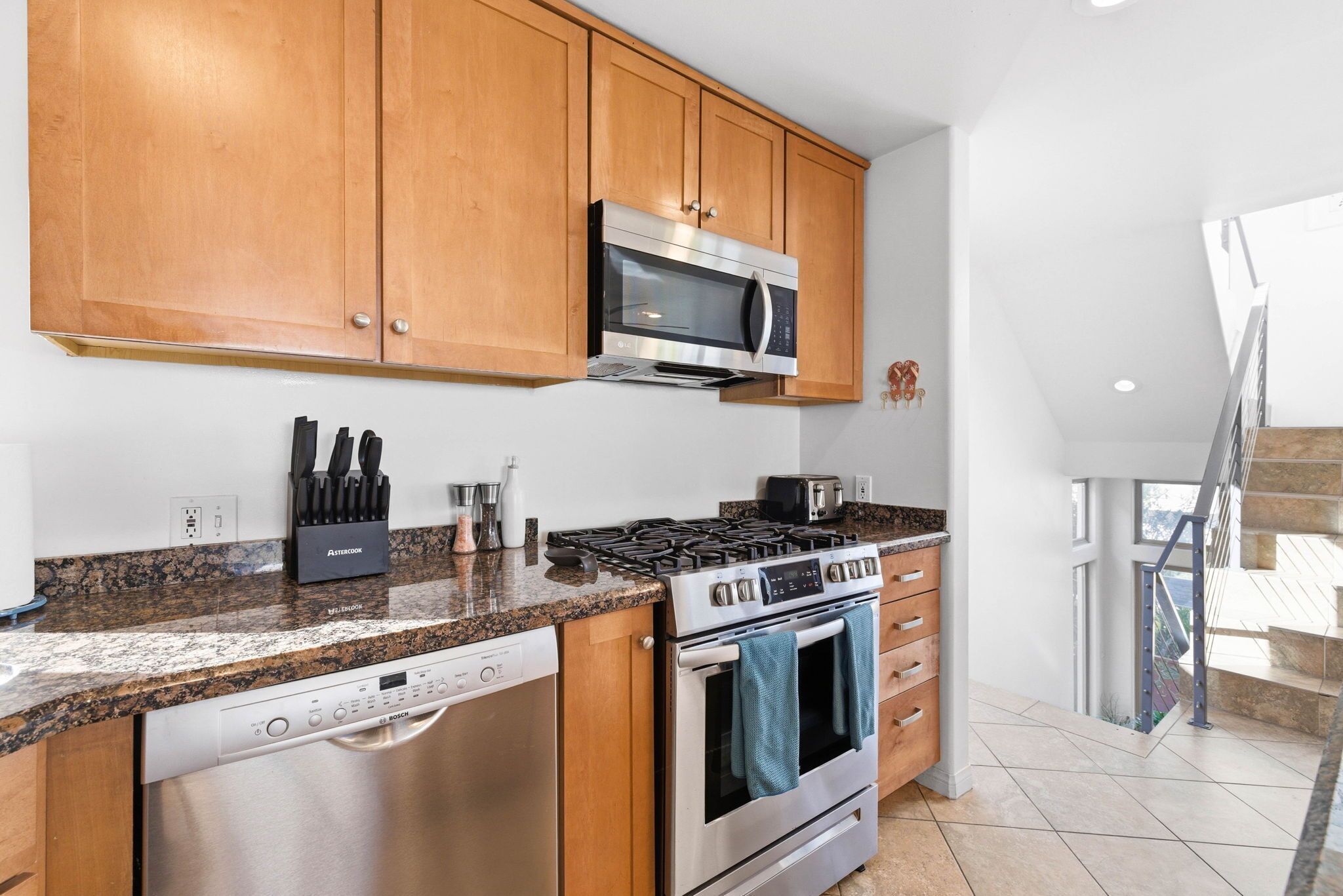 A modern kitchen with wooden cabinets, stainless steel appliances, granite countertops, and a visible staircase leading to an upper level.