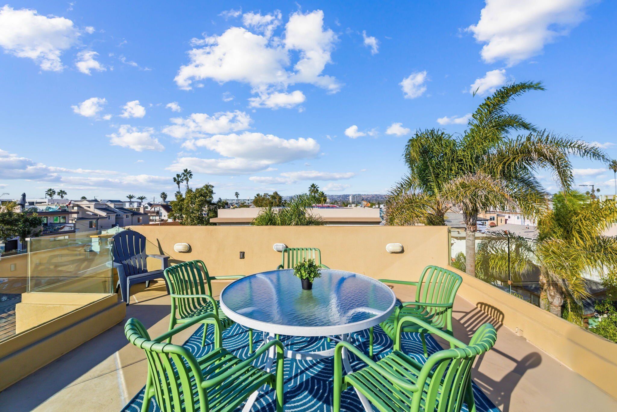 A rooftop terrace with a glass balustrade, green metal chairs, a round table, and a potted plant.