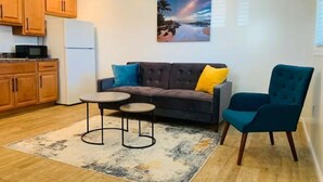 Un espacio habitable compacto con kitchenette, un sofá gris con cojines azules y amarillos, un sillón azul verdoso y un cuadro de una escena de playa.