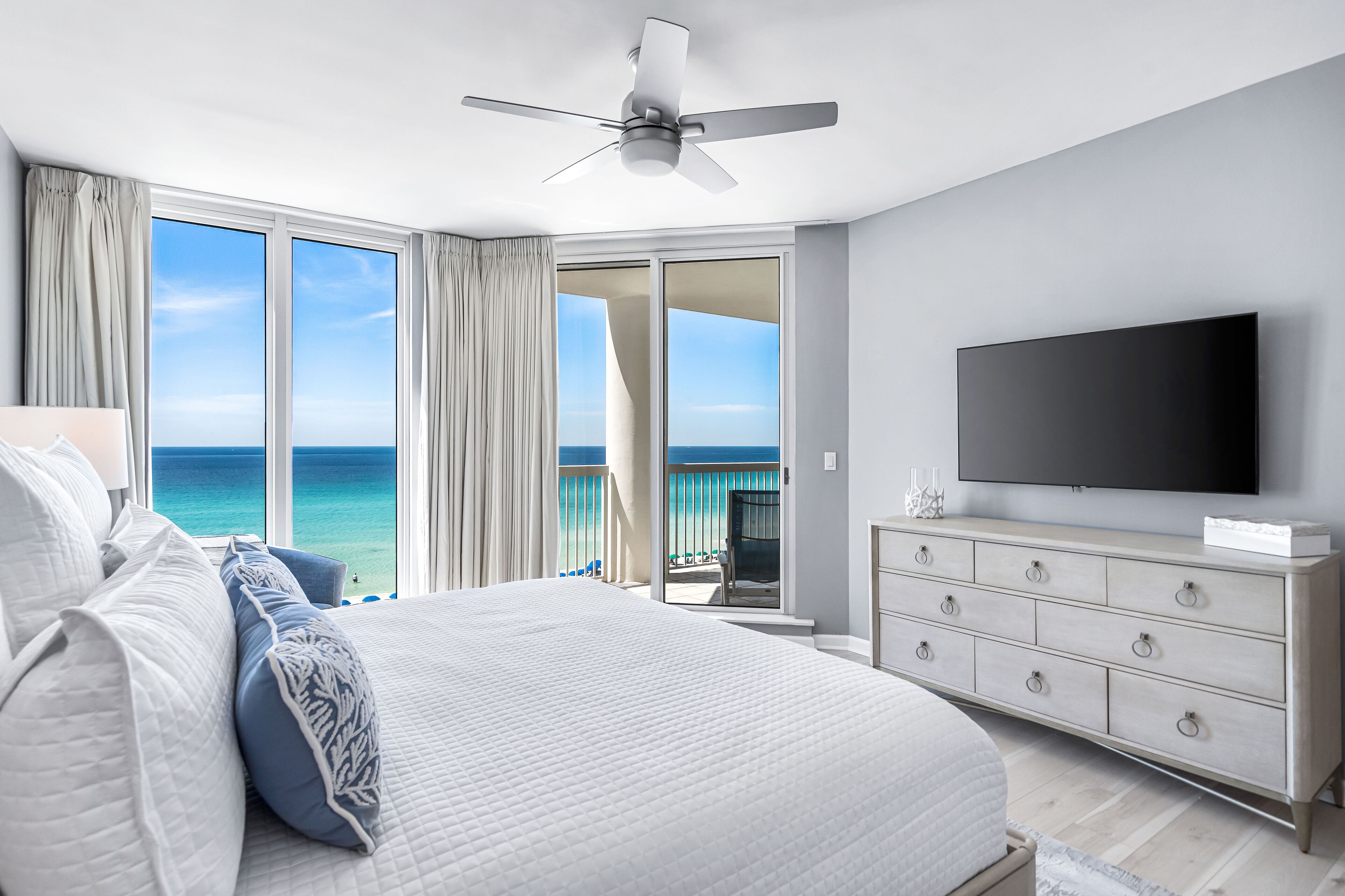 Een slaapkamer met een groot bed, een televisie, een plafondventilator en een balkon met uitzicht op zee.