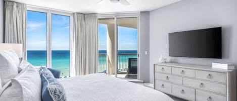 Een slaapkamer met een groot bed, een televisie, een plafondventilator en een balkon met uitzicht op zee.