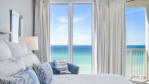Habitación de hotel con una cama grande, una silla y vistas al océano a través de ventanales de piso a techo.