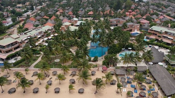 An aerial view of a resort with multiple buildings, palm trees, and a central swimming pool surrounded by beach areas with sun loungers and umbrellas. - Villa Bord de mer Avec Piscine Privée (Saly)