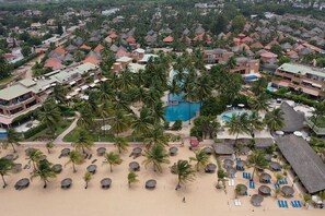 Eine Vogelperspektive zeigt einen Urlaubsort mit mehreren Gebäuden, Palmen und einem zentralen Schwimmbecken, umgeben von Strandbereichen mit Liegestühlen und Sonnenschirmen.