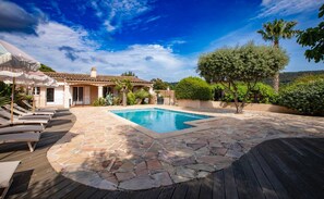 A pool area with stone flooring, lounge chairs, and a house in the background.
