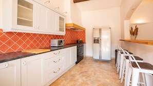 A compact kitchen with white cabinets, a microwave, a stove, and a refrigerator. There are white bar stools and a wooden shelf with decorative items.