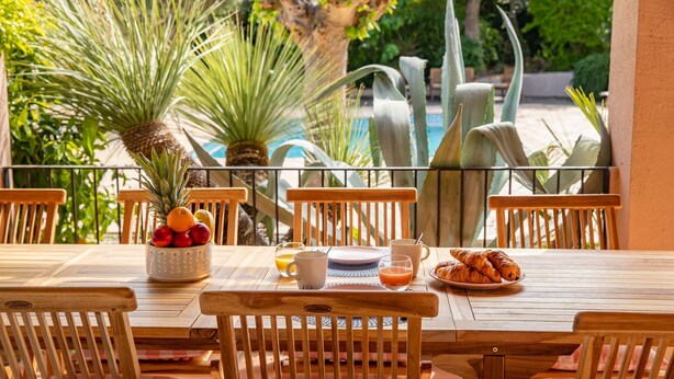 A wooden table set with breakfast items, including fruits, a croissant plate, and a glass of orange juice, in a sunny outdoor setting with a pool and greenery in the background.