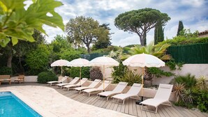 A poolside area with lounge chairs and umbrellas, surrounded by greenery and trees.