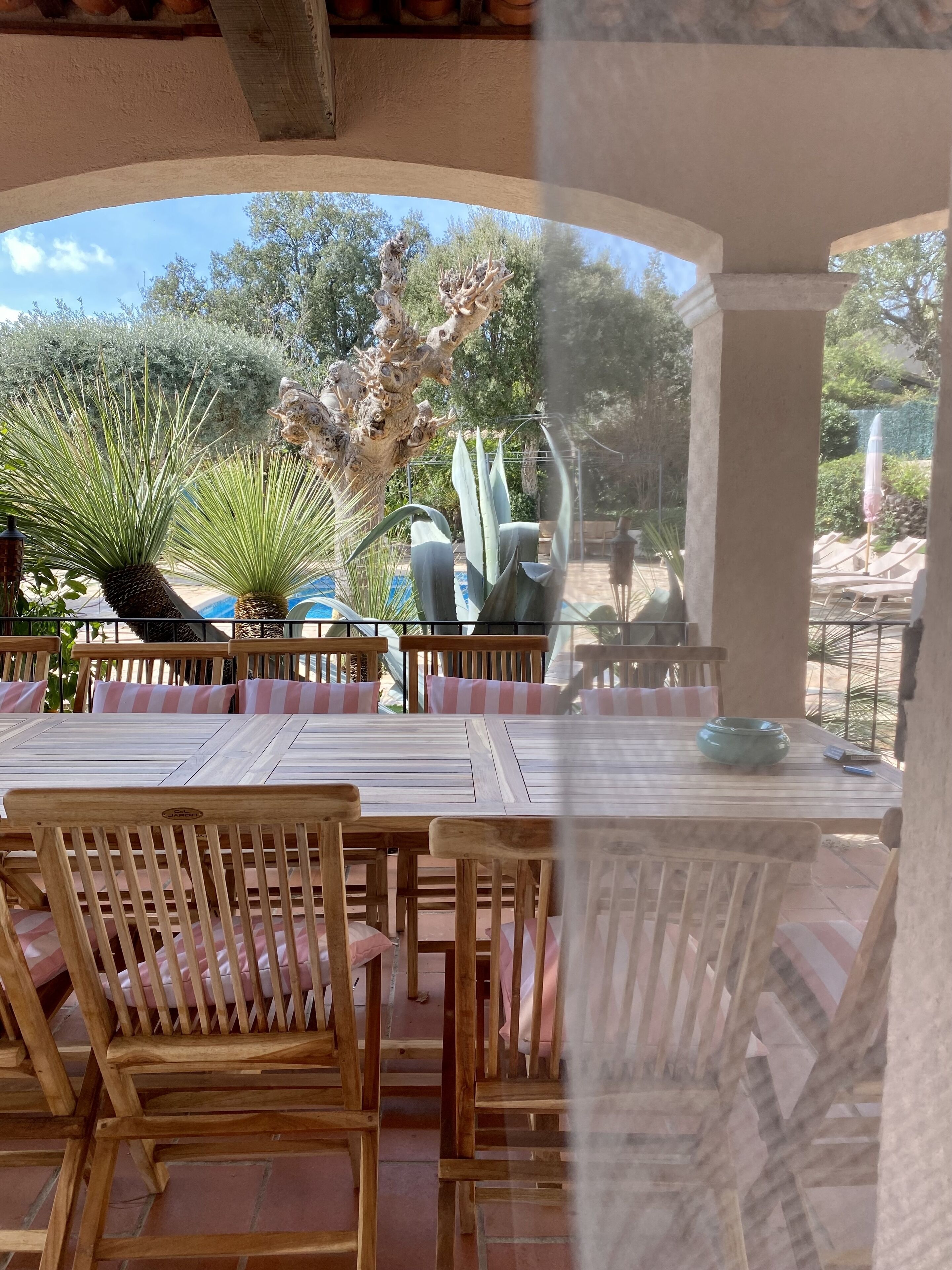 An outdoor patio with wooden tables and chairs, surrounded by various plants and a pool.