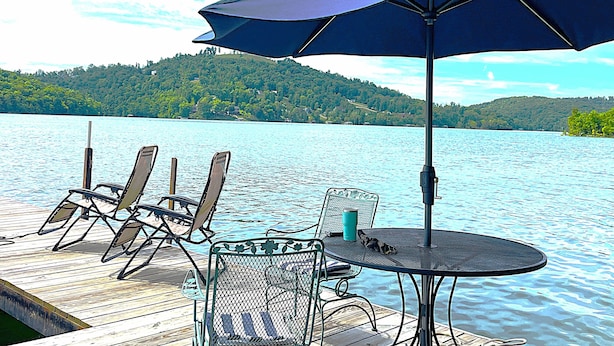 Um deck à beira do lago com mesa, cadeiras e um guarda-sol, com vista para uma colina arborizada.