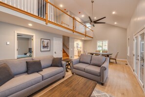 A spacious living area with two grey sofas, a wooden coffee table, and a dining table with chairs. A staircase with a wooden railing leads to an upper level.