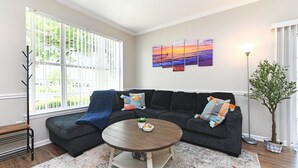 A living room with a sectional sofa, a round coffee table, a large window with blinds, a floor lamp, and a potted plant. - Beautiful Villa in Myrtle Beach within 4 min walk to the beach (Myrtle Beach)
