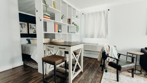 A modern, well-lit room with a wooden floor, a white desk with drawers, a brown leather chair, a black sofa, and a white bookshelf filled with books and decorative items.