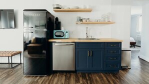 A compact kitchen with a refrigerator, microwave, and dishwasher. There are wooden shelves above the counter and a small seating area with a bench.