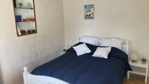 Un dormitorio con cama, cabecera blanca y mesita de noche con lámpara. Hay un cuadro enmarcado en la pared y una estantería con libros y adornos.