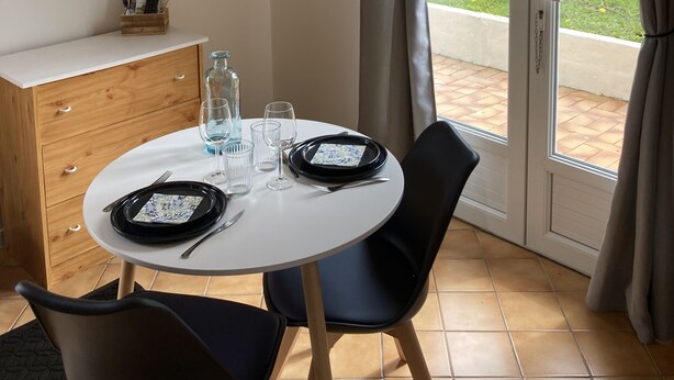 Un comedor con una mesa redonda preparada para dos personas, un armario de madera, un televisor montado en la pared y vistas al jardín a través de la puerta de vidrio.