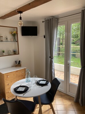 A dining area with a round table set for two, a wooden cabinet, a TV mounted on the wall, and a view of the garden through the glass door. - Gîte de Blay (Blay)