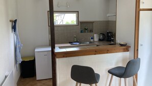 Cocina compacta con un refrigerador pequeño, un lavamanos y un ventanal. Hay dos taburetes de bar y un cubo de basura.
