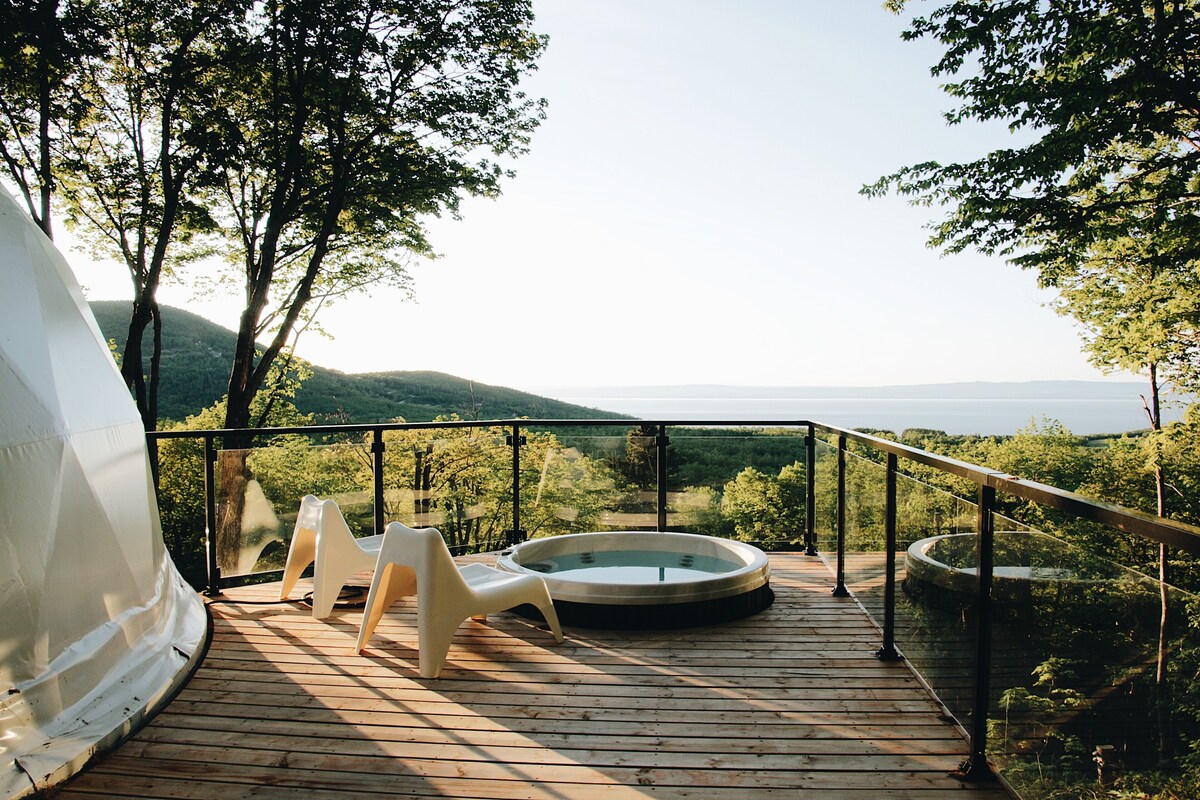 Terraza de madera con un jacuzzi y dos sillas de plástico blancas, con vista a una zona boscosa y un paisaje distante.