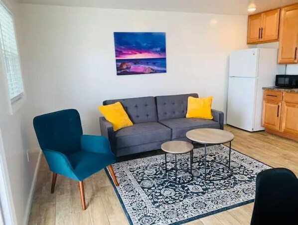 Ein gemütlicher Wohnraum mit grauem Sofa, zwei gelben Kissen, einem smaragdgrünen Sessel und einem kleinen runden Tisch. Ein gemustertes Teppich bedeckt den Boden, und ein Sonnenuntergangsbild hängt an der Wand.