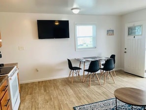 Eine moderne Küche mit Einbauherd, einem Essbereich mit Tisch und Stühlen, einem an der Wand befestigten Flachbildfernseher und einem Fenster für natürliches Licht.