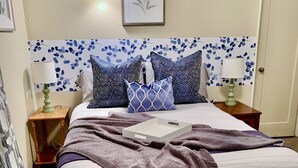 A neatly made bed with a white and purple blanket, flanked by two nightstands with lamps, a wall-mounted light, and a floral-patterned headboard. - Historic District, Downtown, STE 2 (Rapid City)