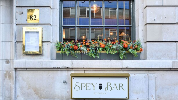 En byggnadsfasad med en skylt som visar texten ”SPEY BAR” och ett fönster med en blomsteruppsättning.