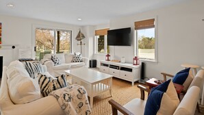Una sala de estar moderna con mobiliario blanco, un televisor de pantalla plana y ventanas amplias.