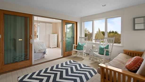 Una habitación amplia con un gran ventanal corredero que da a un balcón, una cama de madera, un sofá blanco y una zona de comedor con sillas blancas.