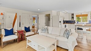 Amplia sala de estar con un sofá blanco, dos sillones y una mesa de centro. Se ve una cocina con salpicadero azul y un comedor con mesa y sillas de madera.