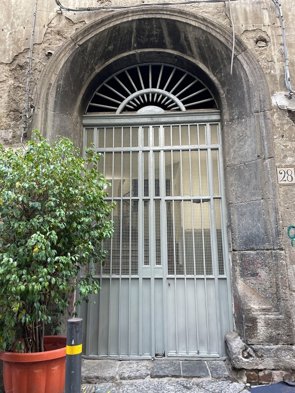 A stone building with a large arched entrance, a metal gate with a sunburst design, and a potted plant in the foreground.