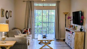Ein Wohnzimmer mit Sofa, Couchtisch, Fernseher und einem Balkon mit Blick auf Grünflächen.