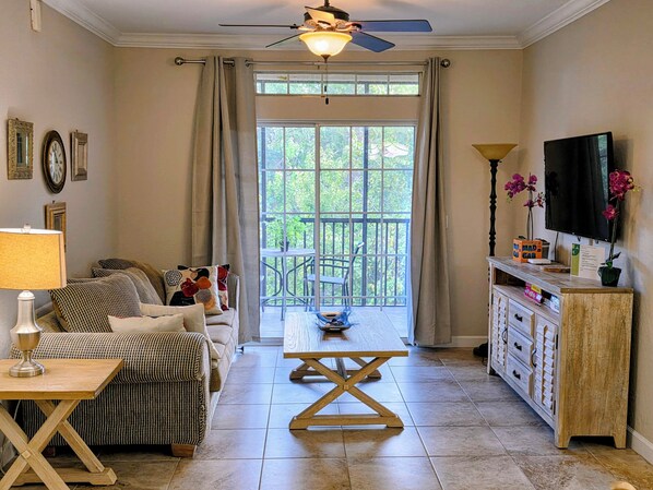 A living room with a sofa, a coffee table, a TV, and a balcony with a view of greenery. - The Sun, Moon, Stars and more!! (Davenport)