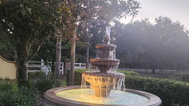 Ein mehrstufiger Steinbrunnen mit herabfließendem Wasser, umgeben von üppigem Grün und Bäumen.