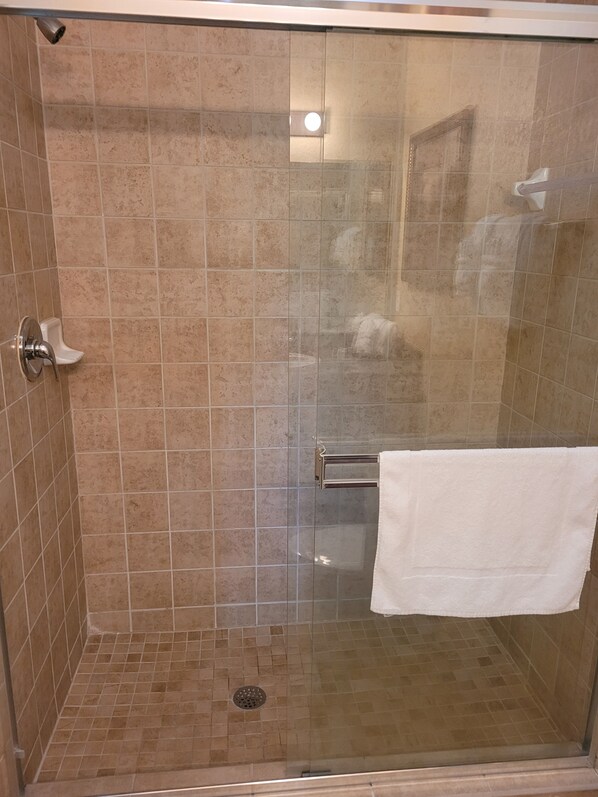 Une cabine de douche fermée par des parois en verre, avec des carreaux de couleur beige, une serviette blanche suspendue à un porte-serviettes et un drain visible.
